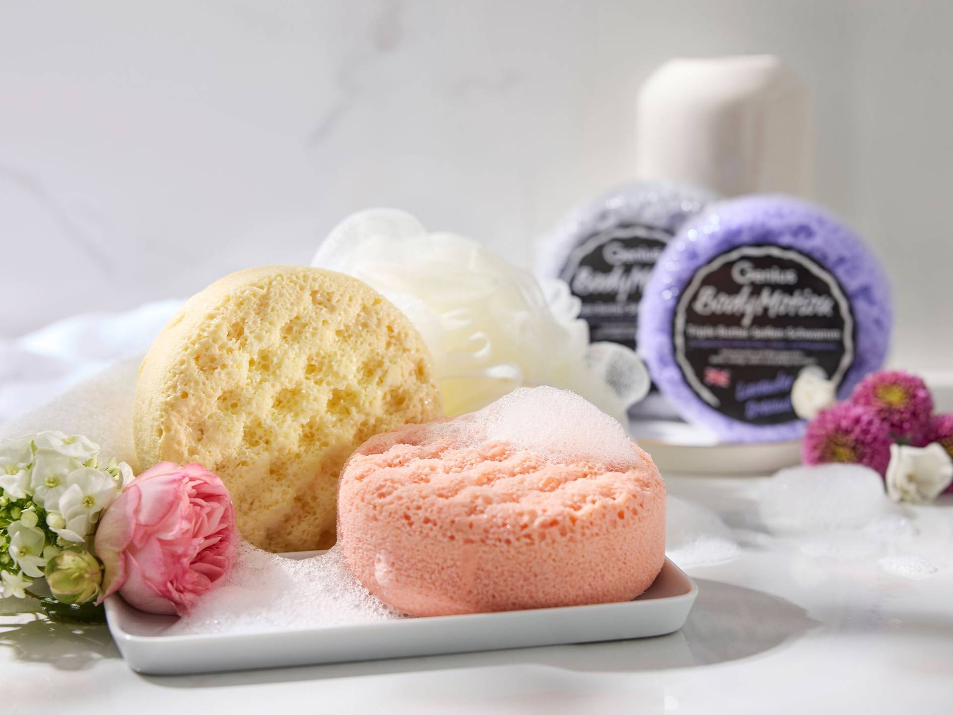 Seife & Schwamm in einem – Pflege neu gedacht Runde Beauty-Schwämme in Gelb und Rosa mit Schaum auf weißem Tablett, umgeben von Blumen und zwei verpackten Schwämmen im Hintergrund
