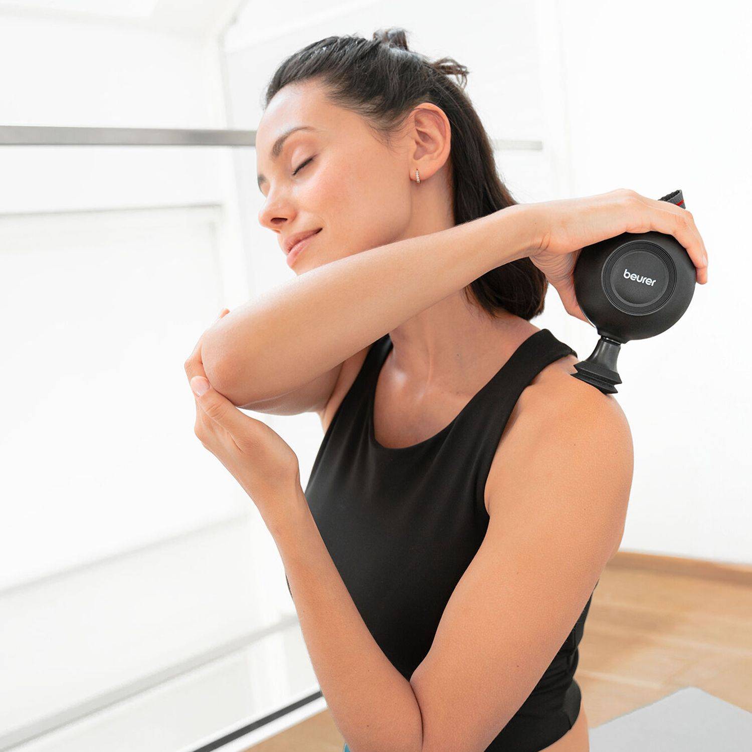 Wohltuende Nackenmassage Frau in schwarzem Sporttop nutzt Massagegerät der Marke Beurer an der rechten Schulter, linker Arm hält Ellenbogen.