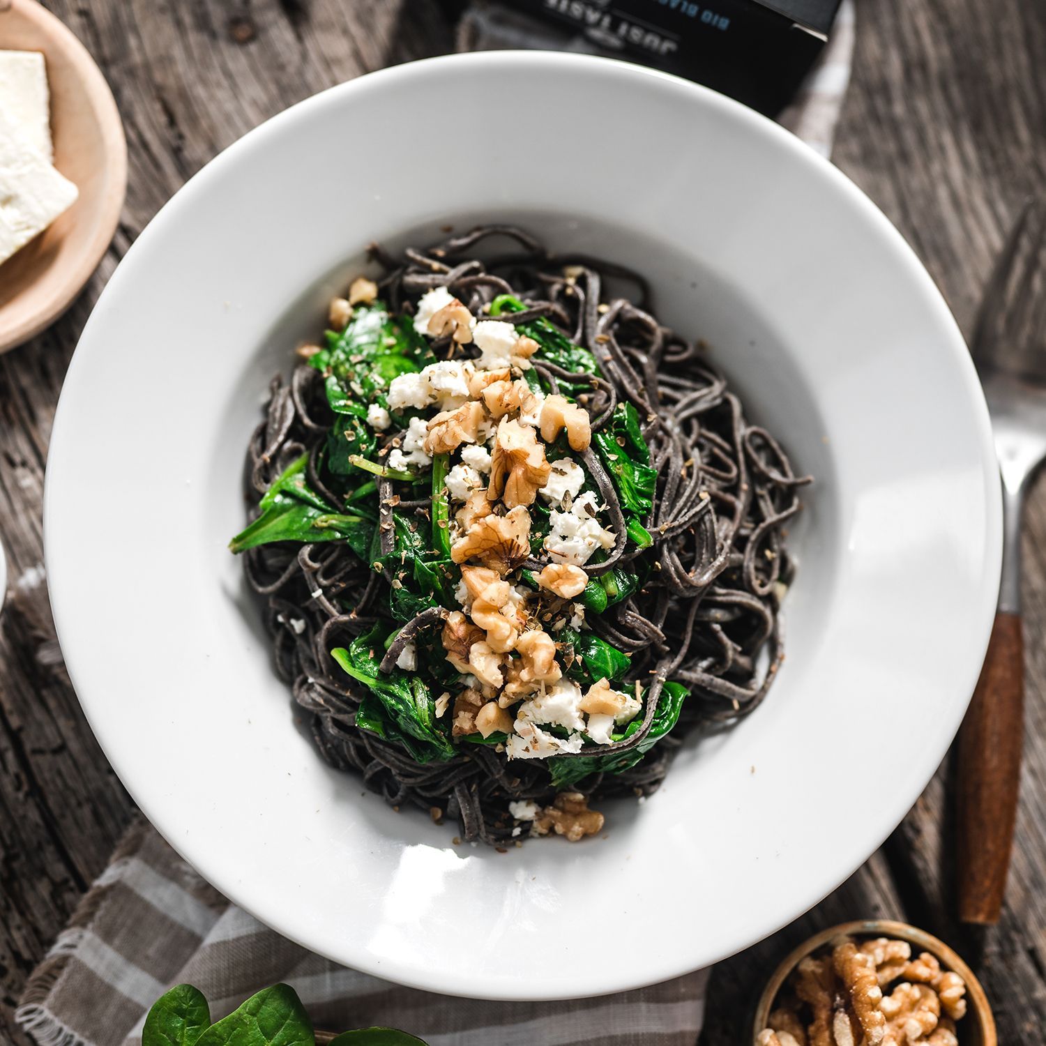 Black Bean Spaghetti mit Spinat, Feta und Walnüssen Schwarze Pasta mit Spinat, Feta und Walnüssen in weißem Teller auf Holztisch, daneben Walnüsse und Verpackung