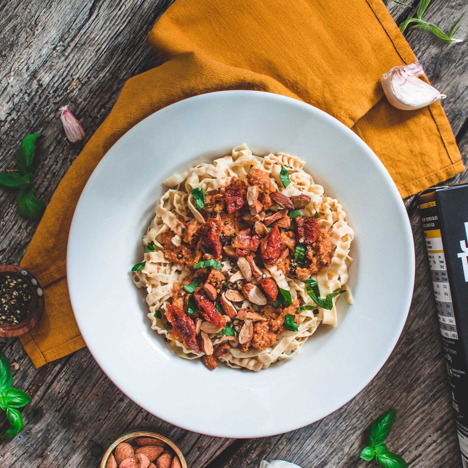 Pasta aus Hülsenfrüchten | Just Taste Weißer Teller mit Pasta, veganer Sojasauce und Kichererbsen, garniert mit Basilikum und Mandeln, auf Holztisch mit Gewürzen und Just Taste Pasta
