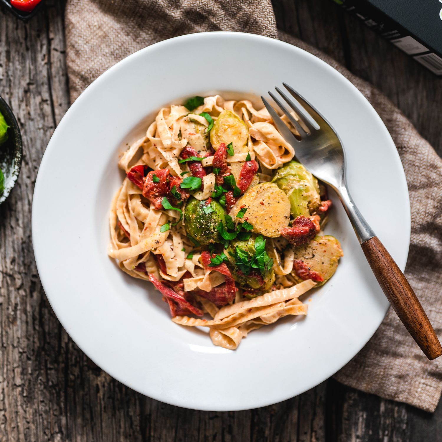 Cremige Rosenkohl-Pasta mit Paprika und Kichererbsen-Fettuccine Teller mit Bandnudeln aus Kichererbsen und Soja, Rosenkohl und Speck, daneben Gabel und Packung der Nudeln