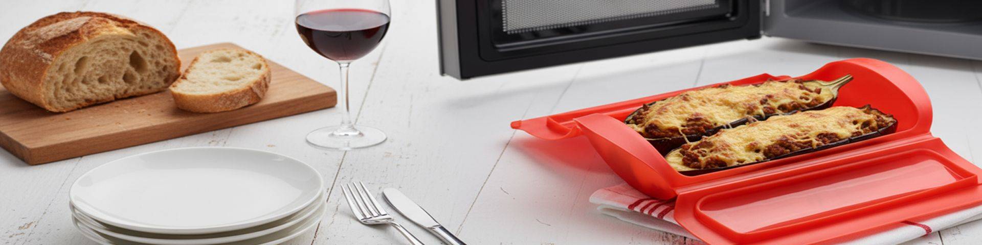 Gefüllte Aubergine in der Dampfbox Zwei längliche, mit Käse überbackene Auberginenhälften auf rotem Backpapier, daneben ein Glas Rotwein, ein Brotmesser, ein Schneidebrett mit Brot und ein leerer weißer Teller mit Besteck.