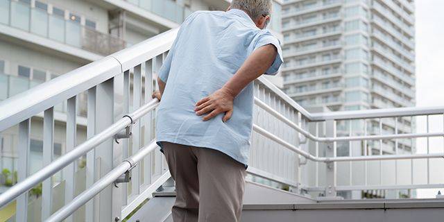 Rückenschmerzen Älterer Mann stützt sich mit einer Hand auf ein Treppengeländer und hält sich mit der anderen Hand den unteren Rücken.