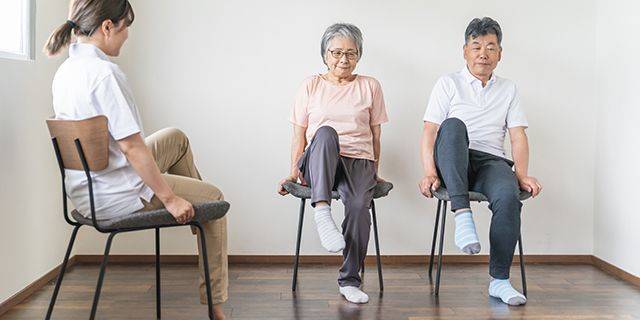 Fitness im Alter Drei Personen sitzen auf Stühlen in einem hellen Raum, zwei heben jeweils ein Bein, die dritte Person sitzt seitlich und beobachtet sie.