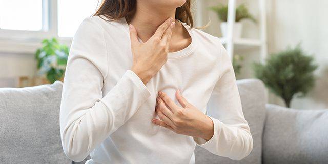 Sodbrennen Frau in weißem Langarmshirt sitzt auf grauem Sofa und hält eine Hand an die Brust, die andere an den Hals.