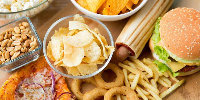 Verschiedene Fast Food Gerichte und Snacks Nahaufnahme von verschiedenen Snacks auf einem Tisch, darunter eine Schüssel Kartoffelchips, ein Hamburger, Pommes frites, Erdnüsse, Nachos und Zwiebelringe.