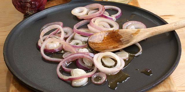 Zwiebeln anbraten Ringscheiben von roten Zwiebeln in einer schwarzen Pfanne mit einem Holzlöffel und etwas Öl.