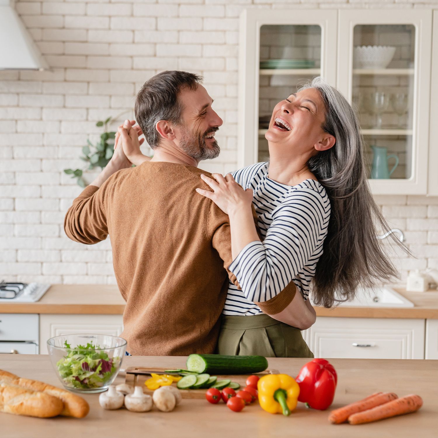 Gesundheitswochen Paar tanzt in einer modernen Küche vor einem Holztresen mit frischem Gemüse und Salat in einer Glasschüssel