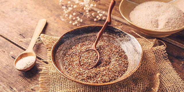 Flohsamenschalen, Kleie und Leinsamen Nahaufnahme von braunen Flohsamenschalen in einer Schale mit einem Holzlöffel, daneben ein weiterer Löffel und eine Schale mit weißem Pulver auf einem groben Stoffuntergrund