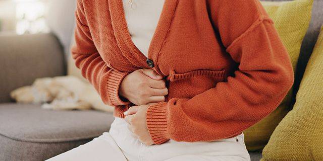 Verstopfung Person im orangen Pullover hält sich mit beiden Händen den Bauch im Sitzen auf einem Sofa