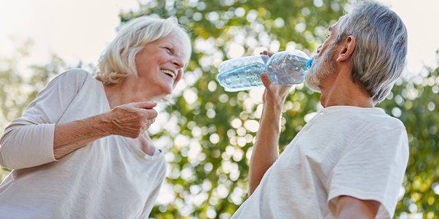 Wasser trinken Älterer Mann trinkt aus einer Wasserflasche im Freien vor unscharfer älterer Frau mit weißem Oberteil